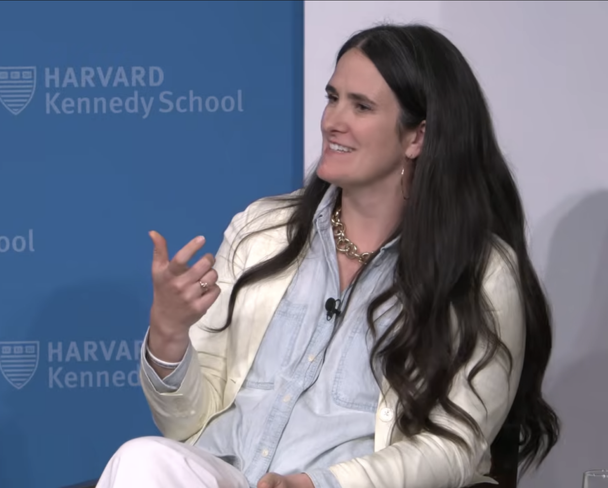 Micaela, a white woman with long, wavy, brown hair, wearing a cream-colored jacket, a blue button-down shirt, and a statement necklace, sits and talks. She is gesturing with her hand and has a microphone clipped to her shirt. Behind her, a blue backdrop with Harvard Kennedy School’s logo patterned on it is visible.